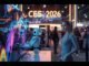 A cinematic, photorealistic, wide-angle shot of a futuristic exhibition hall at CES Las Vegas 2026. In the foreground, a sleek, modern transparent OLED TV displays vibrant colors, positioned next to a friendly-looking Caucasian female humanoid robot helper. In the background, blurred crowds of attendees and glowing neon signage reading "CES 2026" are visible. Caucasian female tech bloggers are actively capturing photos. The aesthetic is cyberpunk yet clean, with warm lighting, high detail, and 8k resolution. The image features cinematic lighting, shallow depth of field, a filmic tone, dynamic composition, and rich color grading.